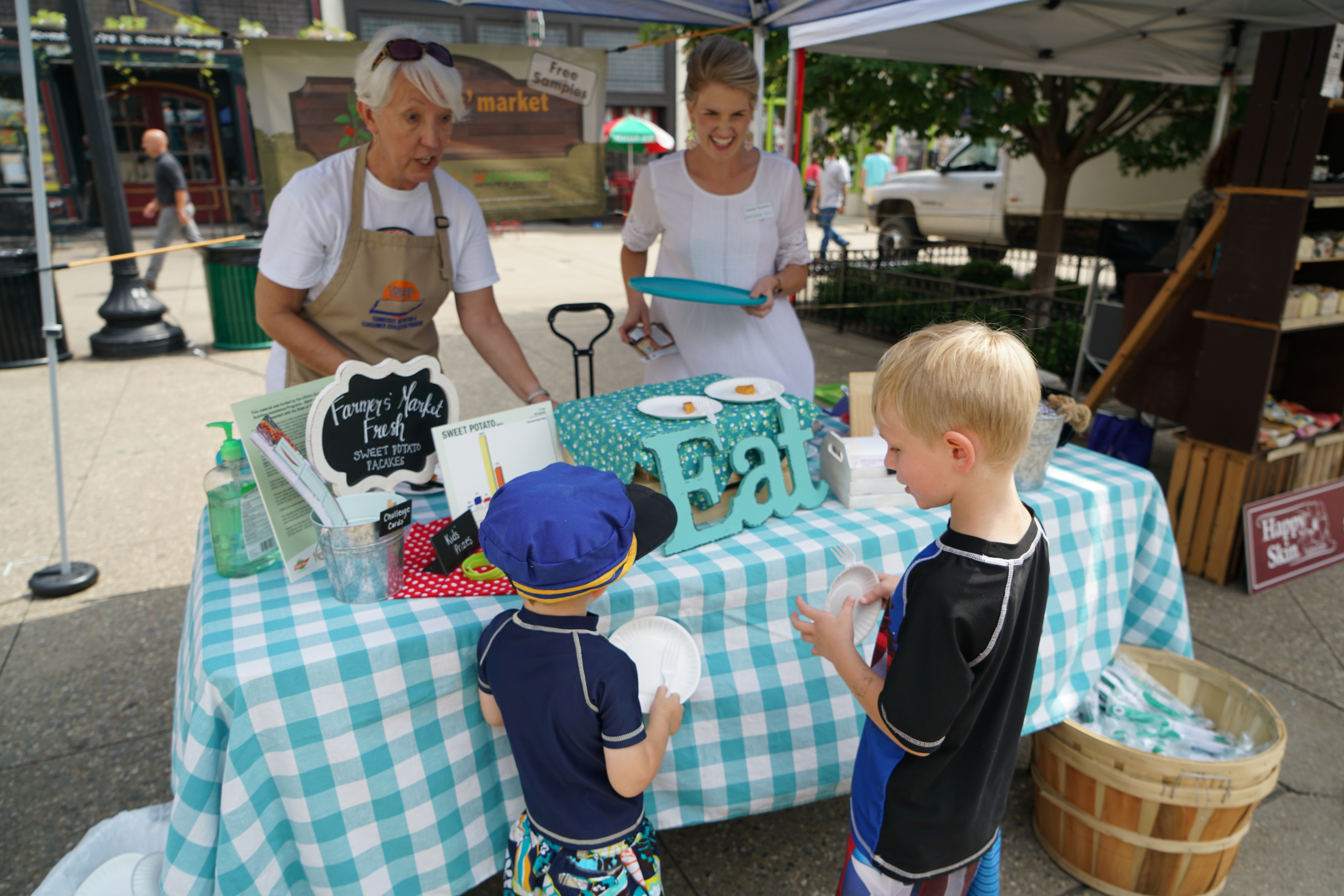 a sample table under a tent at a Farmers Market with two boys trying food