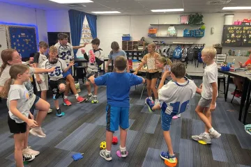 Group of excited 5th graders in althletic clothes in a classroom, standing in a circle, ciking foam dice across the circle to eachother.  